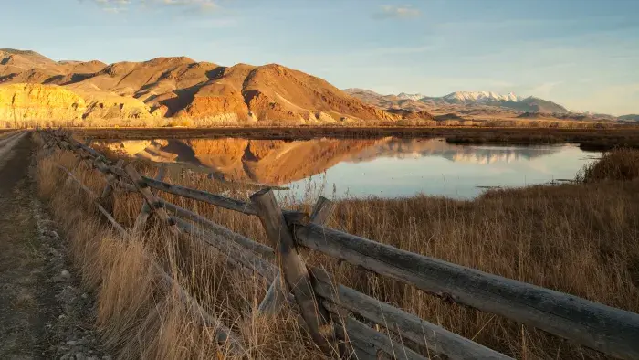 A serene landscape featuring a wooden fence running along a dirt path beside a calm body of water reflecting the surrounding scenery. In the background, golden, sunlit hills rise against a blue sky, with snow-capped mountains visible on the horizon. The foreground includes tall dry grass on both sides of the fence, creating a rustic and tranquil atmosphere.