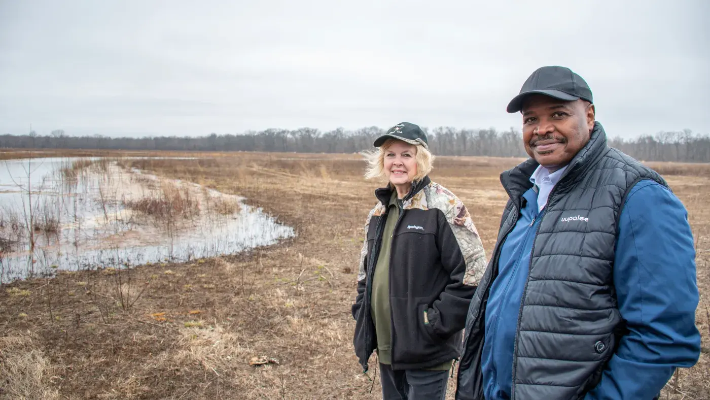 Two people standing by a wetland area in an open field on a cloudy day