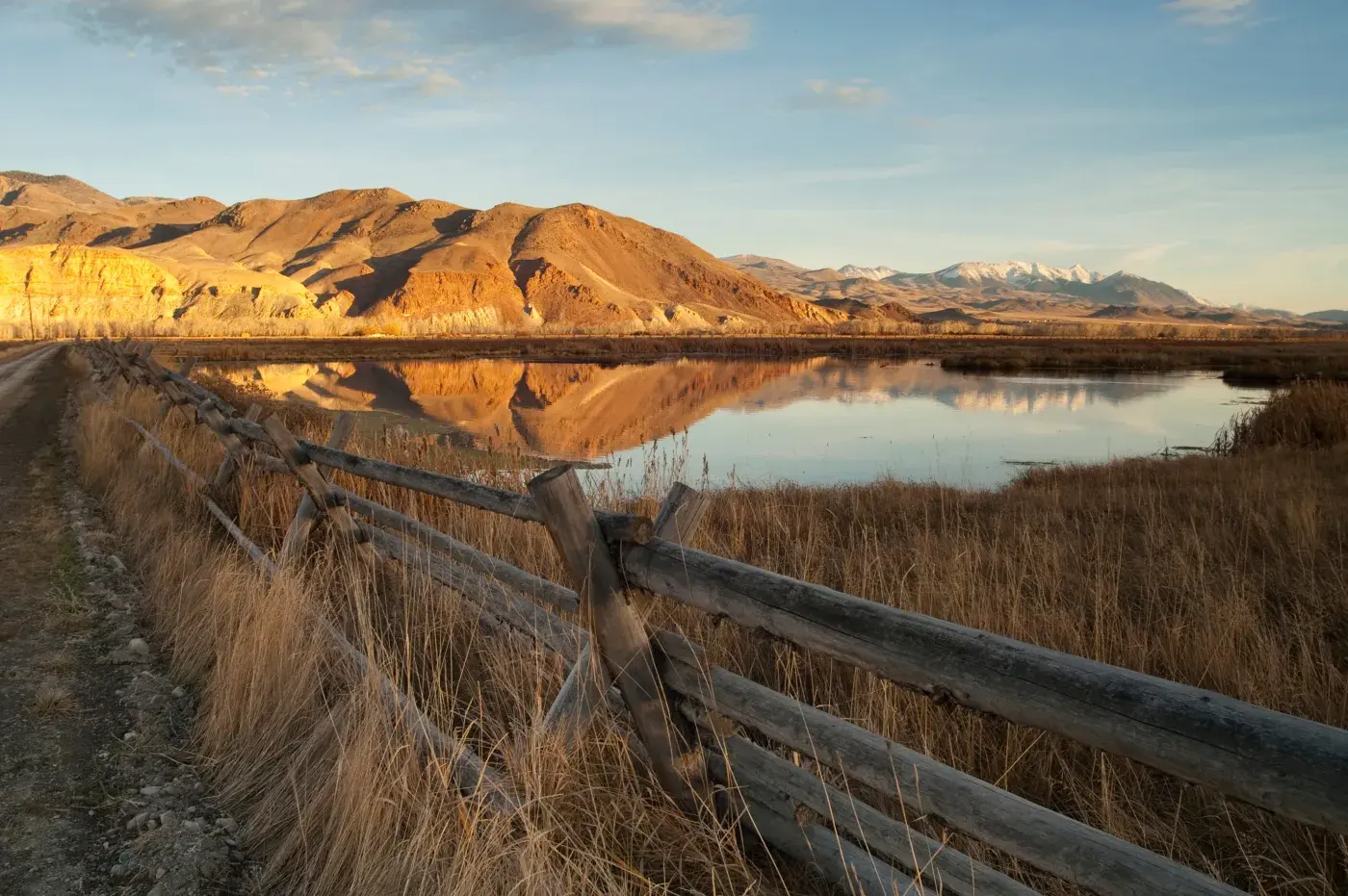 A serene landscape featuring a wooden fence running along a dirt path beside a calm body of water reflecting the surrounding scenery. In the background, golden, sunlit hills rise against a blue sky, with snow-capped mountains visible on the horizon. The foreground includes tall dry grass on both sides of the fence, creating a rustic and tranquil atmosphere.